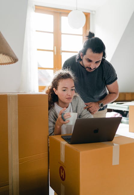 Inside a bright room with natural light coming through a window, a woman and a young girl are engaged in a home relocation process. The woman, with dark hair tied back, is leaning over a cardboard box, observing a laptop placed on top of it. The girl, with curly hair, is sitting beside the box, holding a white mug and focused on the screen. Surrounding them are several packed cardboard boxes, some sealed with packing tape, suggesting they are in the process of packing or unpacking. The room features wooden furniture and a soft hanging light fixture. This scene captures the typical loading, packing, and planning stages of furniture transport and household moves. Highbury Movers, a professional removals company, is involved in overseeing the logistics of this home relocation, with equipment such as boxes and packaging materials visible, emphasizing careful handling and organization during the move.