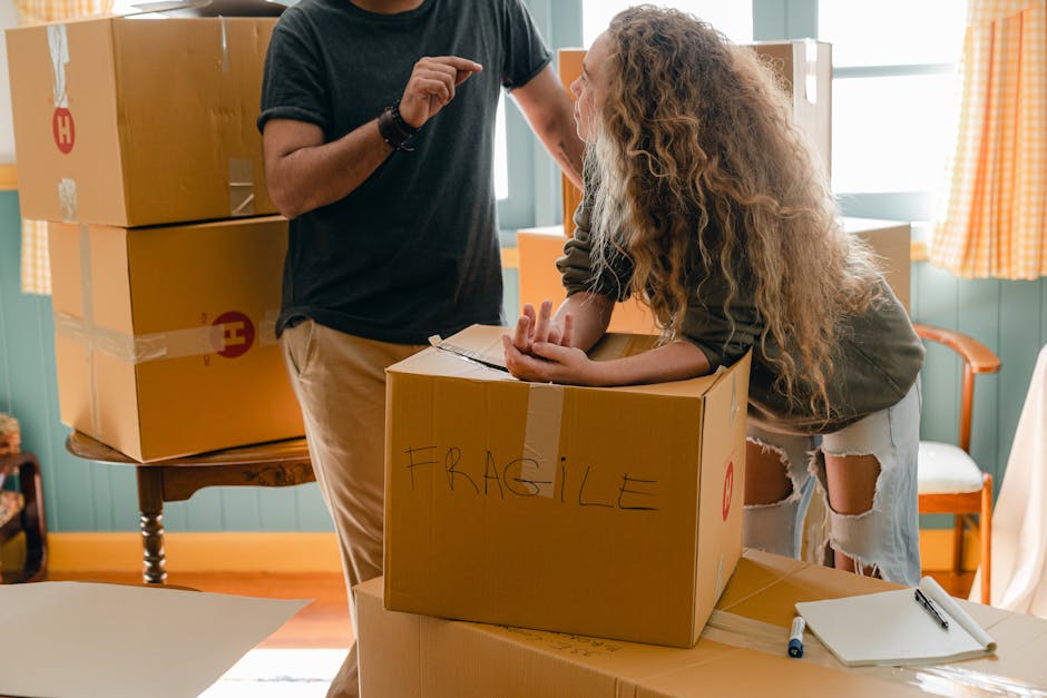 A man wearing a white polo shirt, black cap, and jeans is kneeling on the pavement next to an open cardboard moving box, marking or noting details on a piece of paper or packing list. Several other cardboard boxes, some partially open and filled with packing materials like paper or plastic wrap, are nearby. The scene appears to be set outdoors beside a vehicle or near a loading area, with sunlight illuminating the space, indicating an active home relocation process involving furniture transport and packing. Highbury Movers is conducting a furniture moving or packing and moving service as part of a house removal, with equipment such as packing materials and possibly a van in the background supporting the loading process.