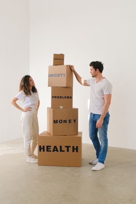 A man and a woman are standing in a spacious, well-lit room with plain white walls and a light-colored floor, engaged in a staging or conceptual activity related to home relocation or emotional preparation. The man, dressed in a white t-shirt and blue jeans, is lifting or placing a small cardboard box labeled 'ANXIETY' on top of a tall stack of four larger cardboard boxes, each labeled with different words: 'HEALTH', 'MONEY', 'PROBLEMS', and 'ANXIETY'. The woman, wearing a white t-shirt and beige trousers, stands to the side with her hands on her hips, observing the process. Nearby, a few empty cardboard boxes are stacked, and the overall setting appears clean and minimalistic, suggesting a staged environment for a move or packing process. The scene subtly reflects themes of emotional baggage and logistics involved in home removals, a service provided by Highbury Movers, as highlighted on their website highburymovers.co.uk.