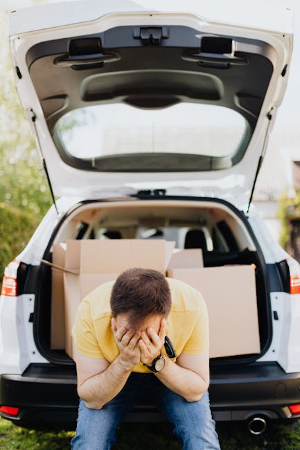 A man wearing a yellow T-shirt and jeans is sitting on the ground in front of an open vehicle trunk during a home relocation process. He is holding his face in his hands, appearing stressed or overwhelmed. Inside the trunk, several cardboard boxes and plastic-wrapped items are neatly packed and arranged, ready for transport. The vehicle, which appears to be a white SUV or van, is situated outdoors in a residential area with greenery and trees visible in the background. The scene captures the challenges involved in furniture transport and packing during a house move, highlighting the importance of professional removals services such as those provided by Highbury Movers, especially to avoid delays and cancellations noted in the page about highbury removals.