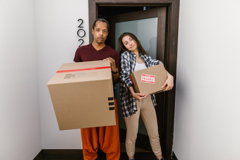 A man and a woman are standing in a spacious, well-lit room with plain white walls and a light-colored floor, engaged in a staging or conceptual activity related to home relocation or emotional preparation. The man, dressed in a white t-shirt and blue jeans, is lifting or placing a small cardboard box labeled 'ANXIETY' on top of a tall stack of four larger cardboard boxes, each labeled with different words: 'HEALTH', 'MONEY', 'PROBLEMS', and 'ANXIETY'. The woman, wearing a white t-shirt and beige trousers, stands to the side with her hands on her hips, observing the process. Nearby, a few empty cardboard boxes are stacked, and the overall setting appears clean and minimalistic, suggesting a staged environment for a move or packing process. The scene subtly reflects themes of emotional baggage and logistics involved in home removals, a service provided by Highbury Movers, as highlighted on their website highburymovers.co.uk.