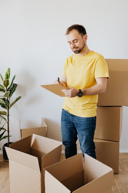 A person wearing a yellow T-shirt and blue jeans is standing indoors next to several large cardboard boxes, some of which are open and empty, while others are sealed with packing tape. They are holding a clipboard in one hand and writing with a black marker in the other, likely making notes related to home relocation tasks. The environment includes a wooden floor, a white wall, and an indoor plant with broad green leaves in a black pot. Behind the person, there are additional packed boxes stacked on top of each other, indicating a packing and moving process is underway as part of furniture transport or home relocation efforts. Highbury Movers, a professional removals company, may be involved in these packing and moving logistics, supporting the transition of belongings during the house removals service.