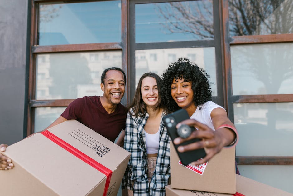 Three individuals, two women and one man, stand outside a modern building with large glass windows during daytime, smiling and taking a selfie together. The woman on the right, with curly black hair, is holding a smartphone and wearing a white shirt with rolled-up sleeves, while the other two hold cardboard boxes wrapped with red straps; the man on the left wears a maroon t-shirt, and the woman in the middle has long brown hair and is dressed in a checkered shirt. The scene depicts a home relocation process involving packing and moving, with moving boxes and friendly expressions. The background includes reflections of buildings in the glass windows, and the setting appears to be in an urban environment suitable for house removals and furniture transport facilitated by Highbury Movers.