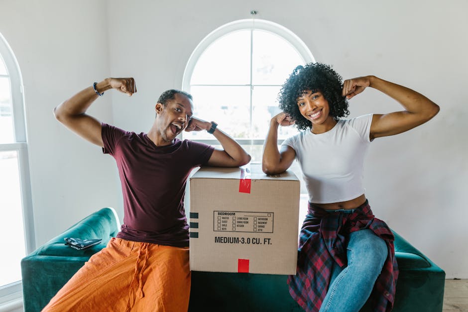A young man and woman sit on a teal sofa inside a bright room with white walls and an arched window, smiling and flexing their arms to show strength during a home relocation process. The man, wearing an orange pair of trousers and a maroon T-shirt, and the woman, dressed in light blue jeans and a white cropped T-shirt with a red plaid shirt tied around her waist, are positioned beside a medium-sized cardboard box labeled with checkboxes and dimensions indicating it contains bedroom items. The box is placed on the sofa between them, suggesting they are preparing for packing or unpacking furniture and household items. The room appears well-lit with natural daylight streaming through the window. Visible behind them are some loose magazines or documents on the sofa, and the overall scene depicts an organised and positive move-in or move-out situation. This image represents the packing and moving stage of a house relocation, with the involvement of Highbury Movers, a professional removals company specializing in residential moves.