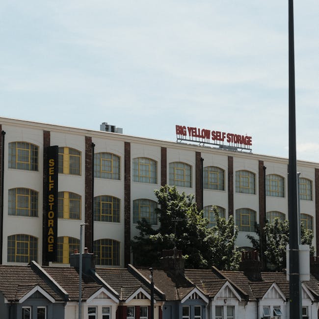 The image depicts a multi-storey building with a large sign reading 'BIG YELLOW SELF STORAGE' on the rooftop, indicating a self-storage facility. The building has a white façade accented with brick columns and large, arched windows with yellow frames. A vertical sign on the side of the building reads 'SELF STORAGE' in yellow letters on a black background. In front of the building, there are residential-style rooftops and small chimneys, with a few trees partially obscuring the view of the lower floors. The scene is set during daylight with a partly cloudy sky overhead. This setting is relevant to house removals, as self-storage facilities like this are often used in home relocation and furniture transport processes, which are typically coordinated by companies such as Highbury Movers for efficient packing, movement, and storage during property transfers.