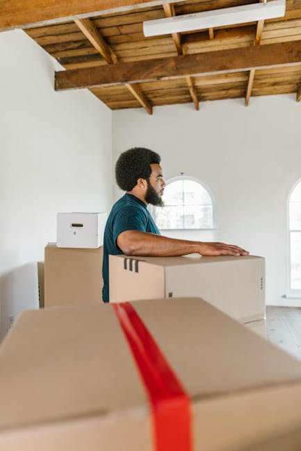 A man with curly black hair and a beard, wearing a dark teal T-shirt, is standing inside an unfinished room with white walls, wooden ceiling beams, and large arched windows. He is resting his arms on top of a large cardboard box, part of a moving process involving house removals. Several other cardboard boxes, some sealed with red packing tape, are stacked around him, indicating packing and moving preparations. The boxes vary in size and are placed on the light wooden floor, with additional boxes on a piece of furniture or a table nearby. The natural daylight coming through the windows illuminates the space, highlighting the ongoing home relocation activity. The scene reflects the logistics of furniture transport and packing in a professional moving service environment, with Highbury Movers occasionally involved in the process.